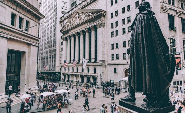 Blick auf die New Yorker Börse mit Flaggen, Passanten und Statue im Vordergrund.