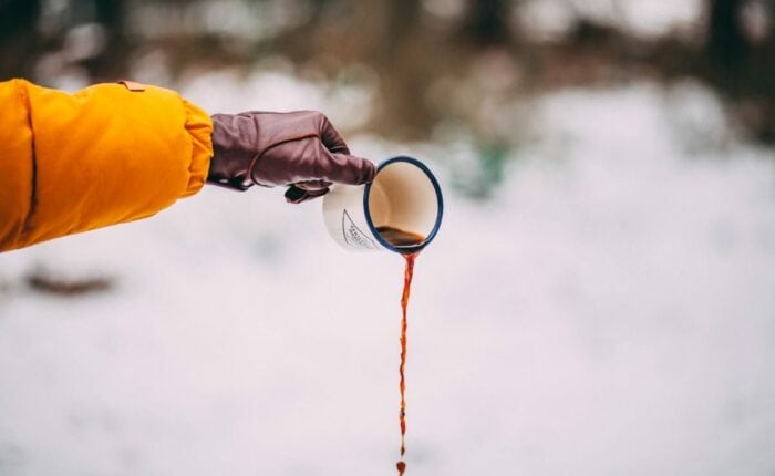 Person gießt Kaffee aus einem Emaille-Becher in den Schnee.