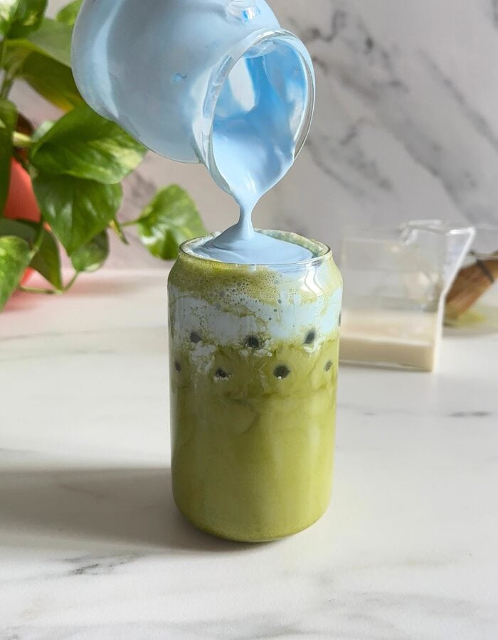 spirulina matcha latte being made, spirulina cold foam being poured, a plant, a milk jug and chasen in the background, on a marble background