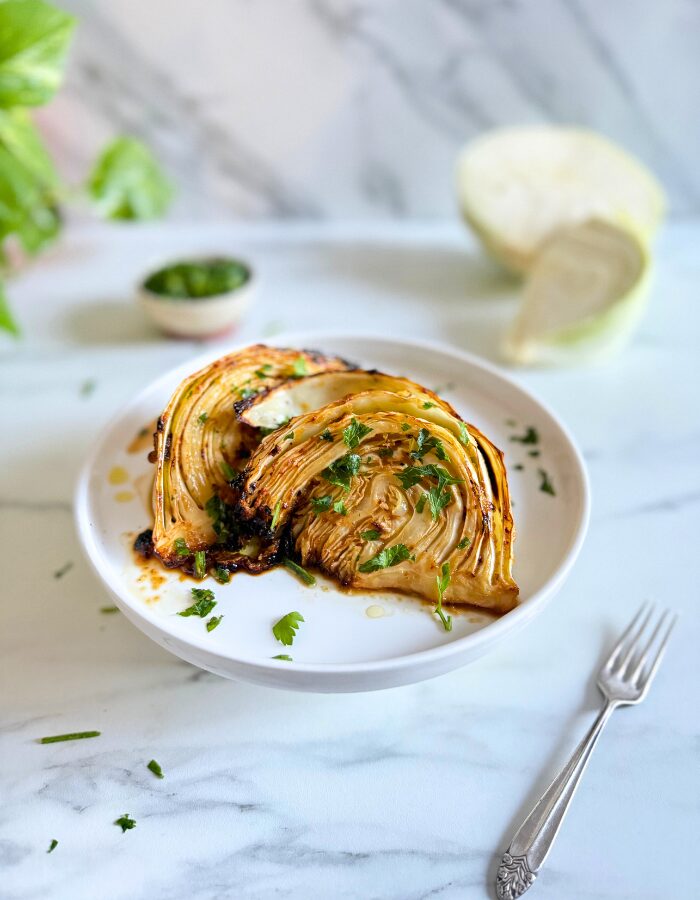 air fryer cabbage wedges on a plate, a fork beside, behind a bowl with cut parsley, a cabbage, and a plant, on a marble background
