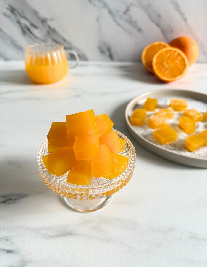 vegan fruit jellies on a cake stand, more fruit jellies on a plate in the back with sugar, oranges and a glass of orange juice, on a marble background