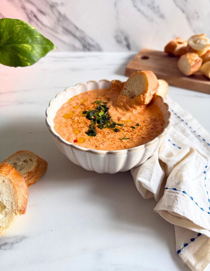Feta Paprika Dip - Easy & Quick Dip Recipe 4 paprika dip in a bowl with a slice of bread, more bread on a wooden board behind and some on the side, a towel, a plant, on a marble background