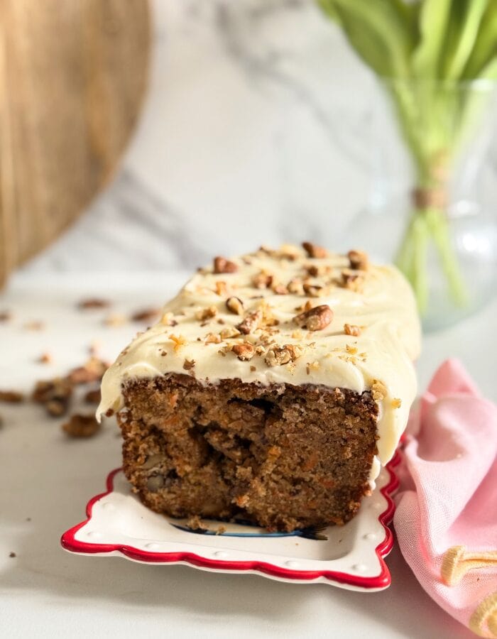 carrot cake pound cake on a plate, pin towel on the side, in the back a vase with tulips and wooden boards, walnuts around, on marble background