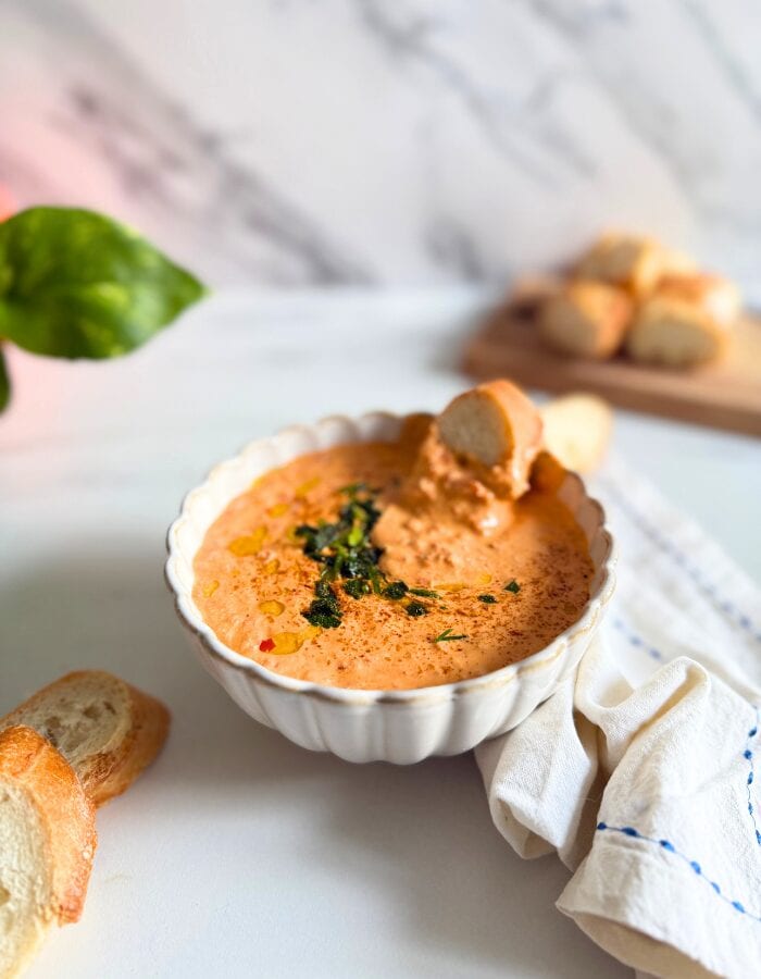 Feta Paprika Dip - Easy & Quick Dip Recipe 6 feta paprika dip in a bowl with a slice of bread, more bread on a wooden board behind and some on the side, a towel, a plant, on a marble background