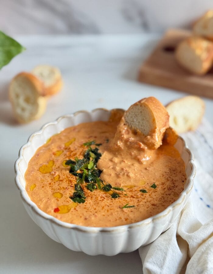 Feta Paprika Dip - Easy & Quick Dip Recipe 1 paprika dip in a bowl with a slice of bread, more bread on a wooden board behind and some on the side, a towel, a plant, on a marble background