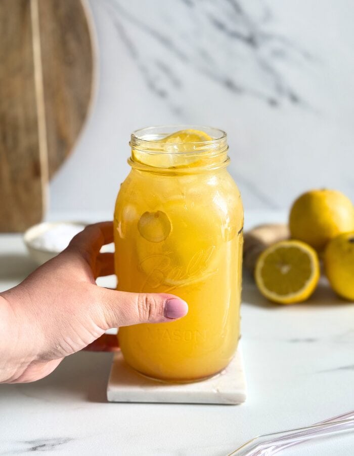 hand holding a jug of natures gatorade on a marble coaster, straws on front, lemons and ginger in the back with a bowl of salt
