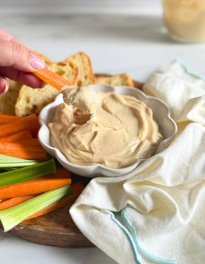 hand holding a carrot dipping into a bowl of miso mayonnaise, on a wooden board with more veggies cut on the side, slices of bread, towel and a jar of mayo in the back