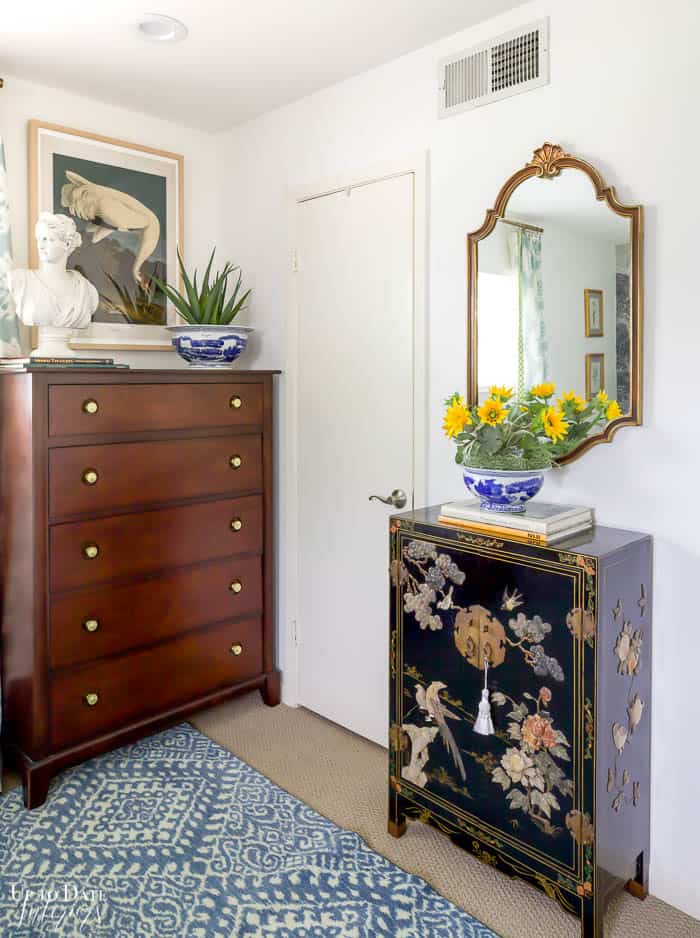 Corner of bedroom with tall brown dresser and black asian cabinet decorated with plants, books, and a bust. Wall art and gold mirror on the walls.