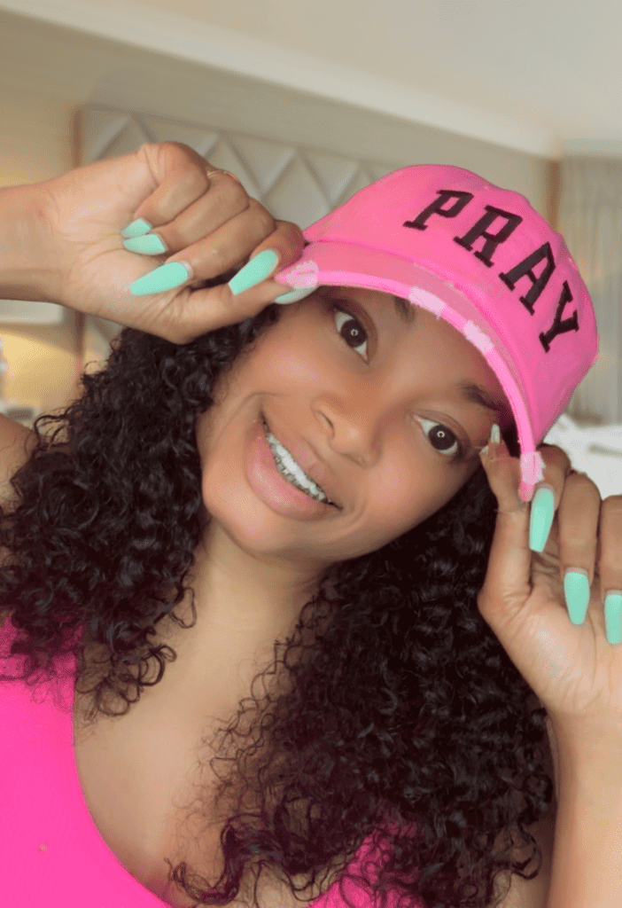 A cheerful woman with curly hair in a hot pink hat with black lettering that says PRAY and a hot pink shirt, happy that God answers prayers. The background is a cozy looking atmosphere.