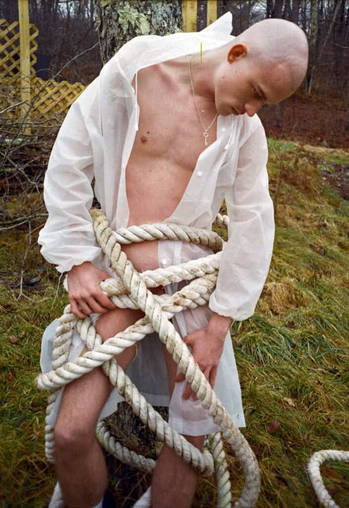 A bald young man in an open white sheer shirt wrapped in thick coiled rope outdoors, head bowed, a key pendant visible at his chest — photograph by Slava Mogutin