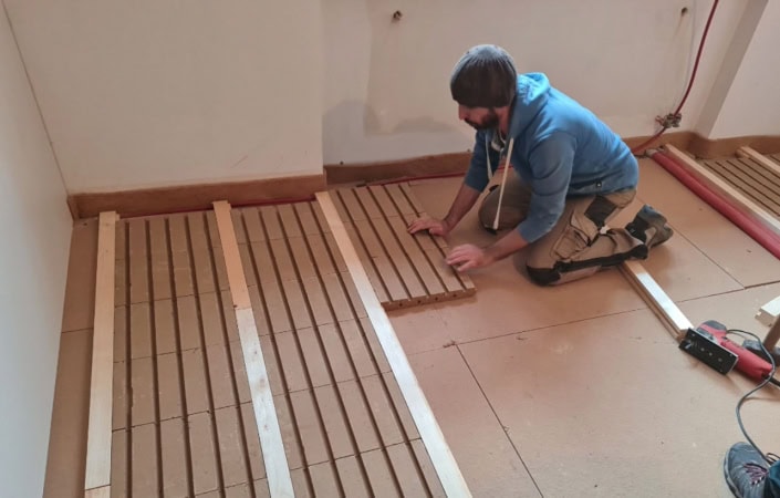 Handwerker kniet und verlegt Lethtermo‑Bodenheizung aus Holzlatten