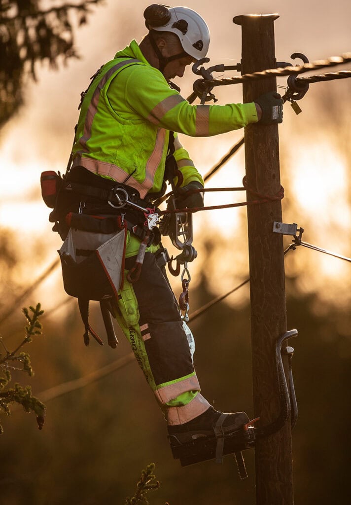 Linjemontör från Norrtälje Energi i säkerhetssele och hjälm arbetar uppe i en stolpe med elledningar i skogsmiljö. Personen ler mot kameran och är omgiven av höst.