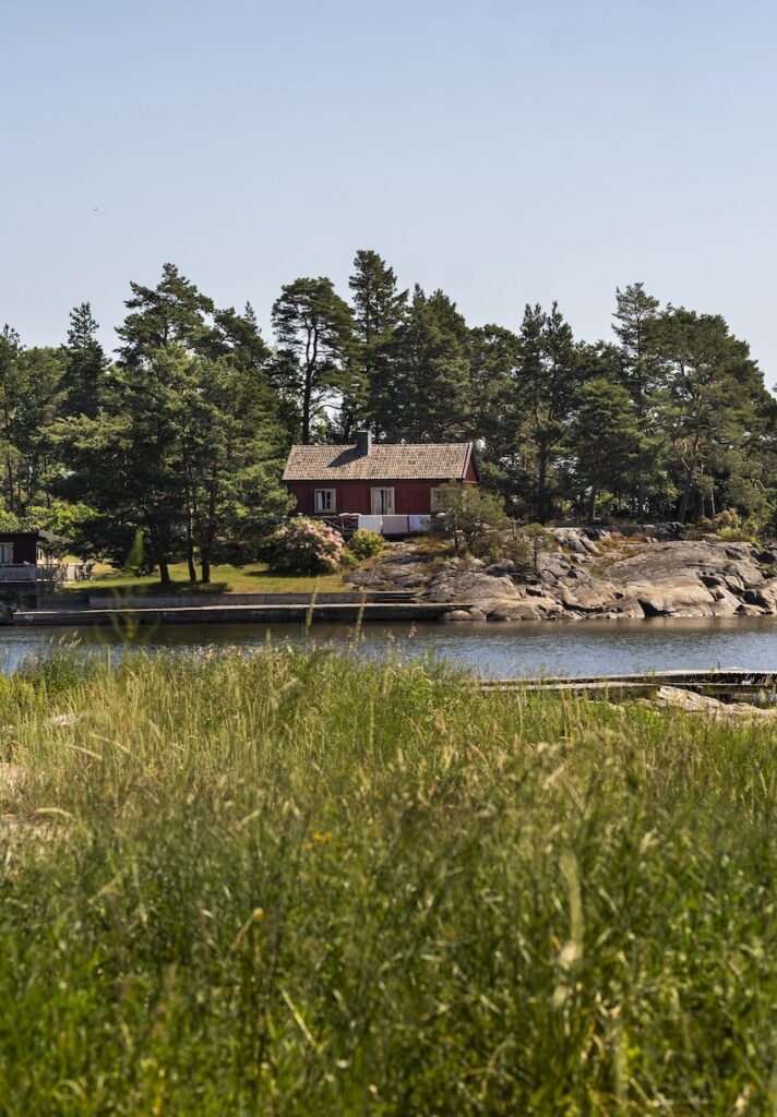 Foto i svensk skärgård på sommaren, från grönska mot en liten röd sommarstuga.
