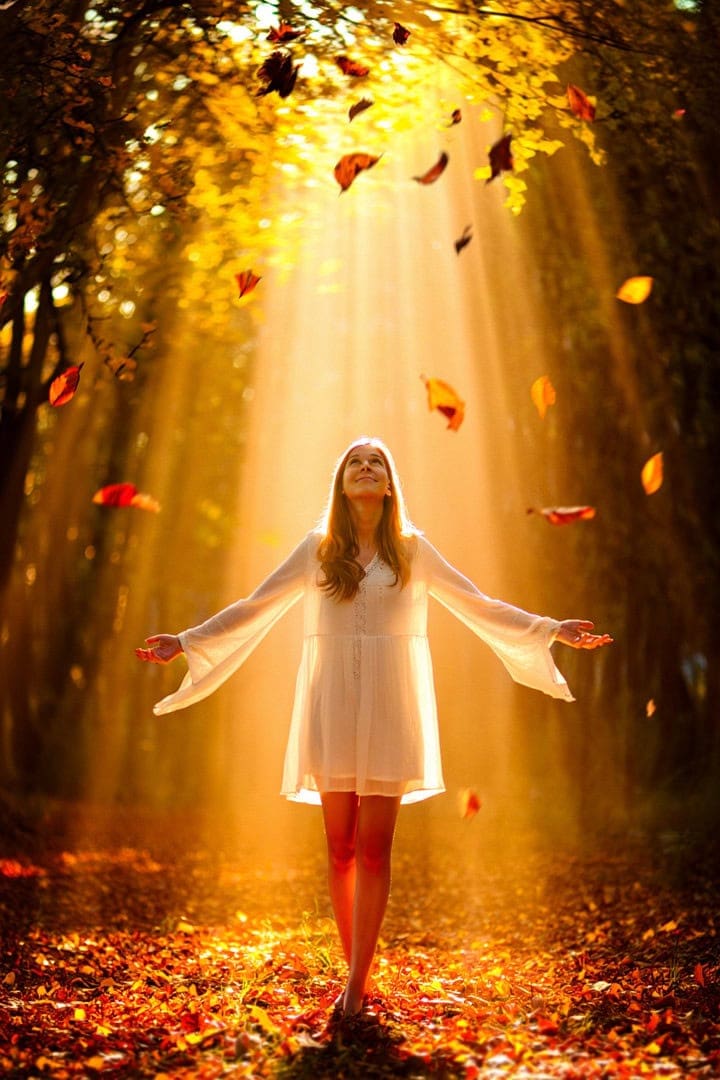 Woman in white dress enjoying autumn forest with falling leaves and golden sunlight.