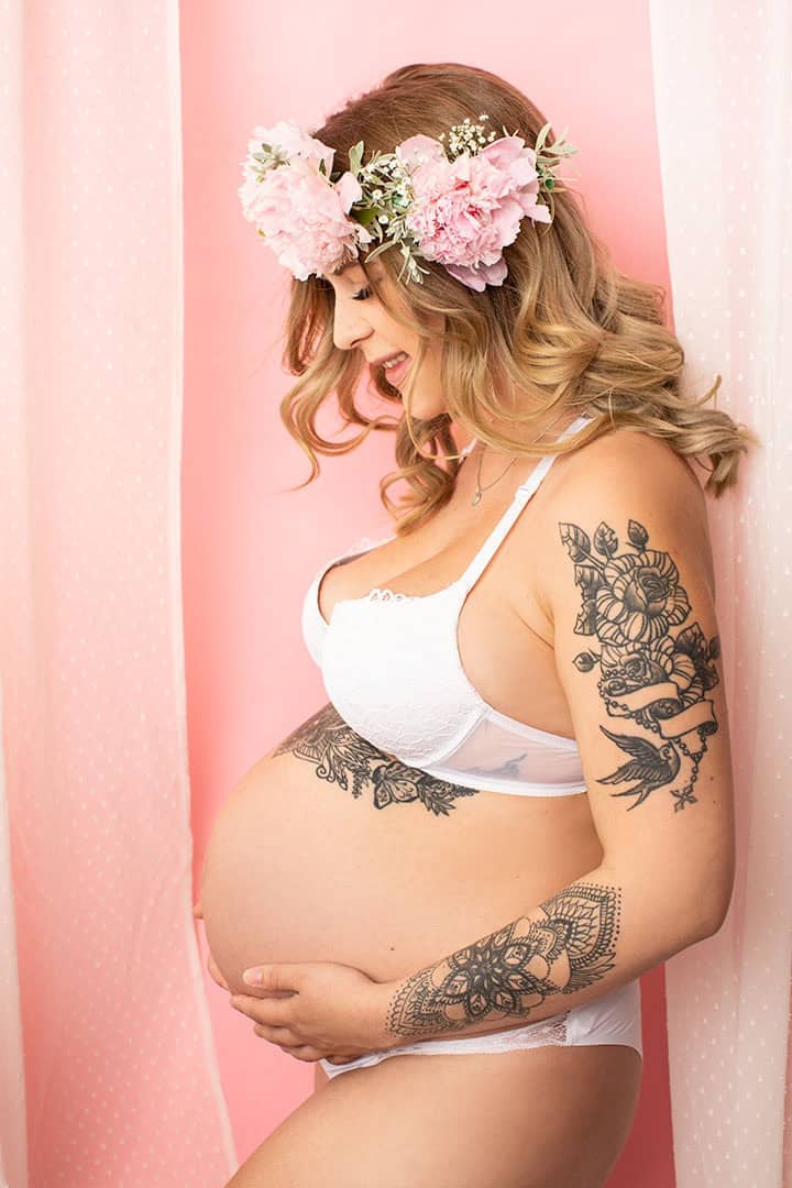 Fødselsfotografering av gravid kvinne med blomsterkrans, smilende og kretsende bakgrunn i rosa.