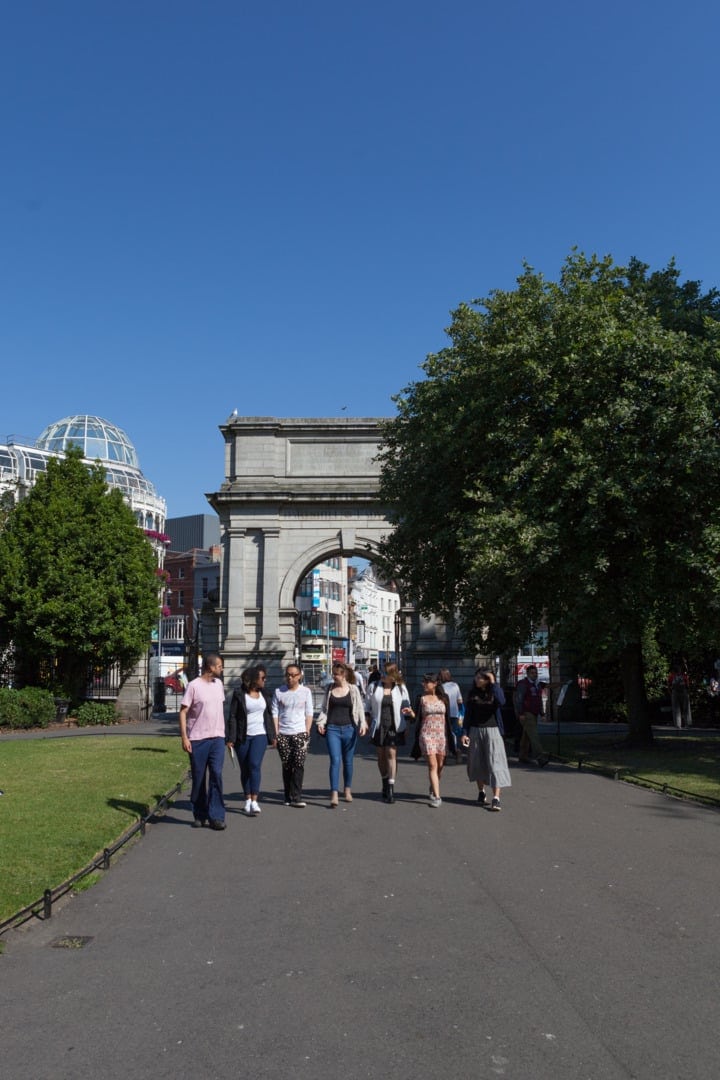 Eine Gruppe von Menschen spaziert durch den St. Stephen’s Green Park in Dublin mit dem Fusiliers’ Arch im Hintergrund.