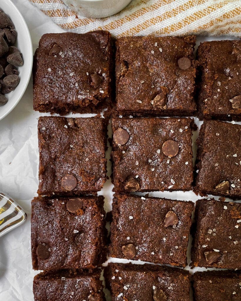 Top view of a grid of brownies laying on a wax paper and yellow/white striped kitchen towel backdrop. A white bowl of chocolate chips sits in the upper left-hand corner.