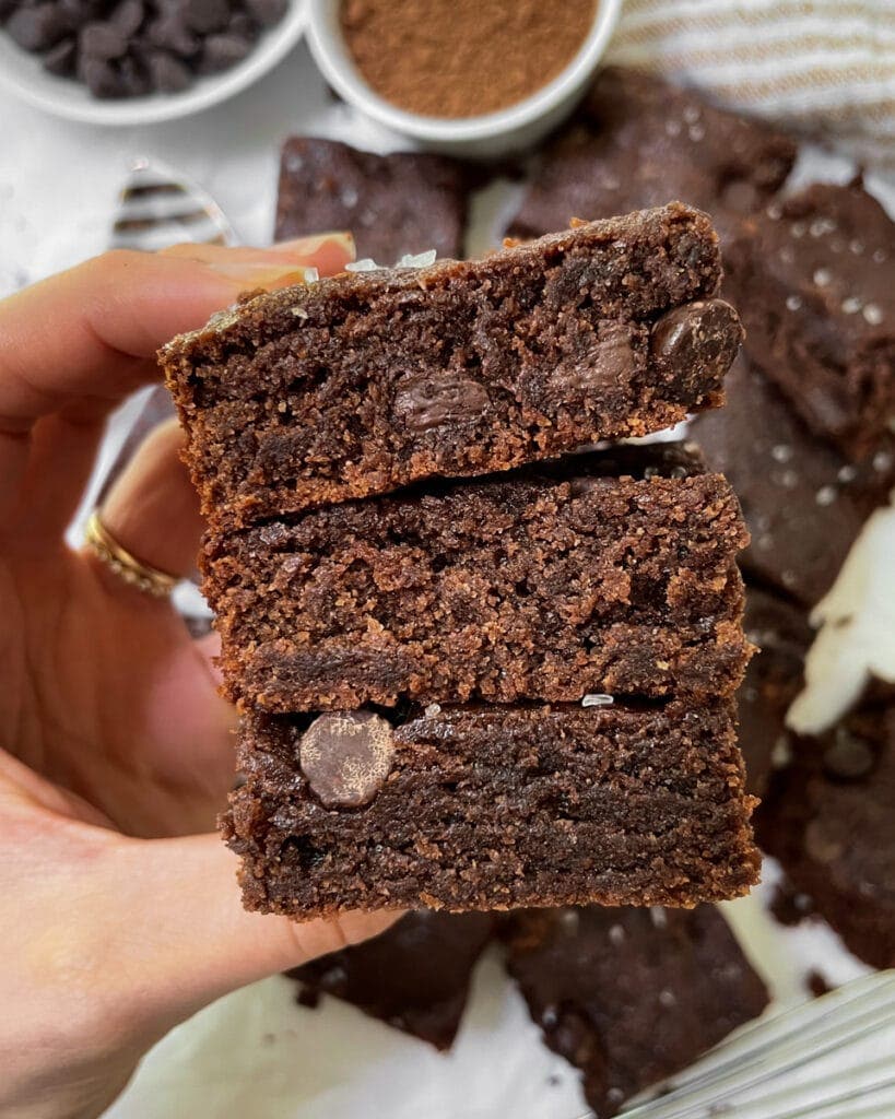 A hand holds three triple-stacked tahini brownies in the foreground. Background shows top view of blurry plate of brownies and two bowls of ingredients.