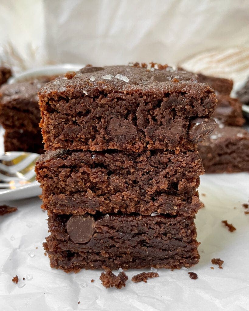 Front shot of triple stack of Tahini Brownies in foreground. Blurred background of more brownies on a wax paper platform.