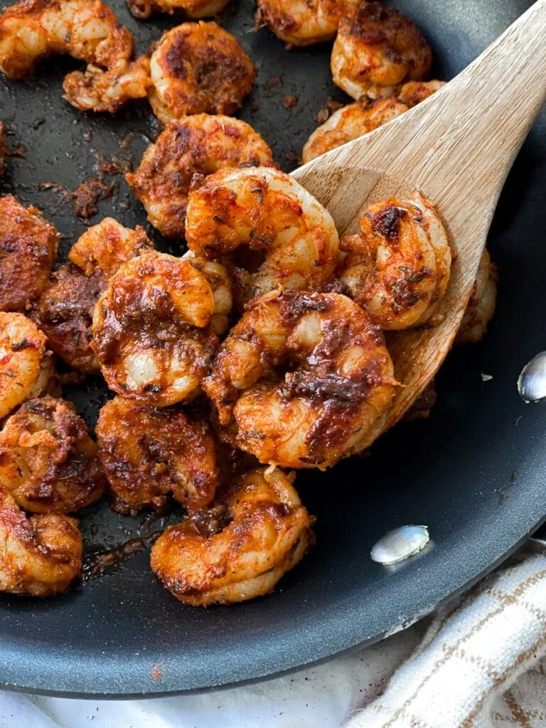close up of blackened shrimp in a skillet next to hand towel with wooden spoon