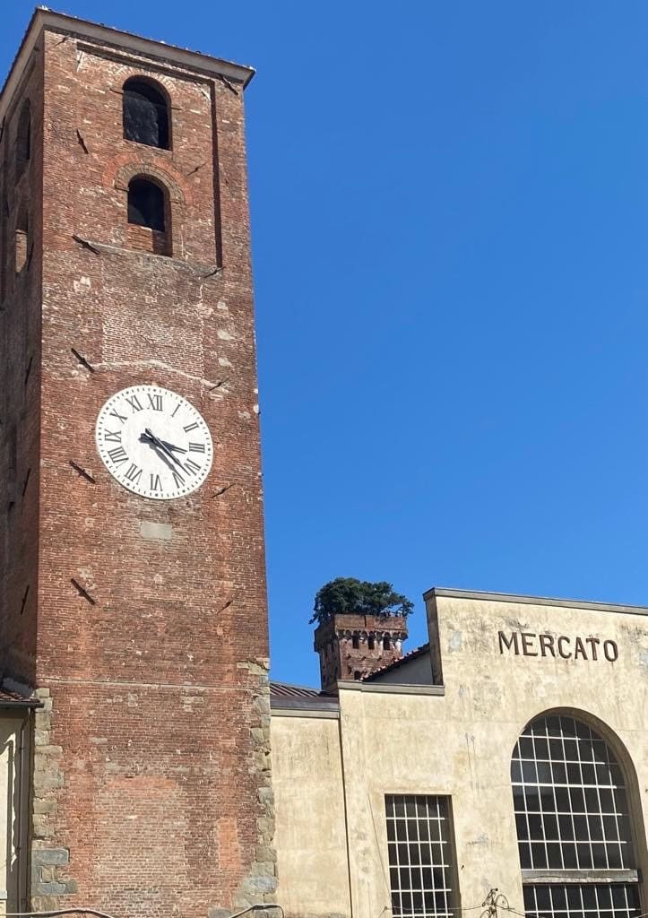 Historischer Backsteinturm mit Uhr und dem Schriftzug „MERCATO“ auf einem Gebäude, blauer Himmel im Hintergrund.