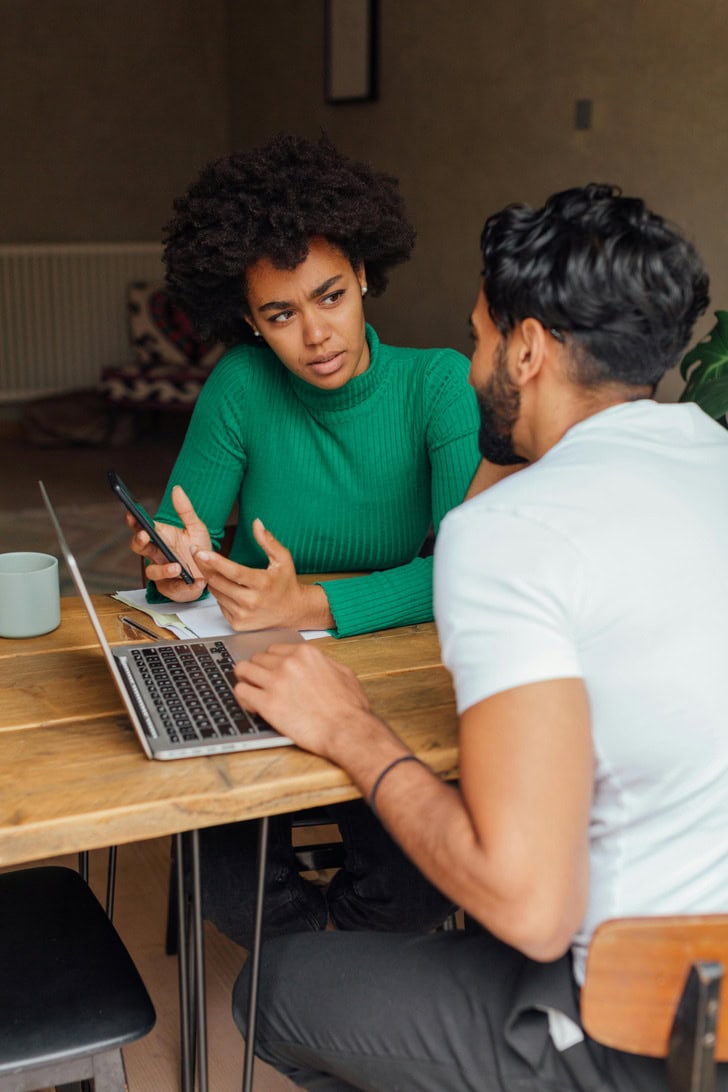 A woman with a phone talking seriously to a man working on a laptop at a wooden table.