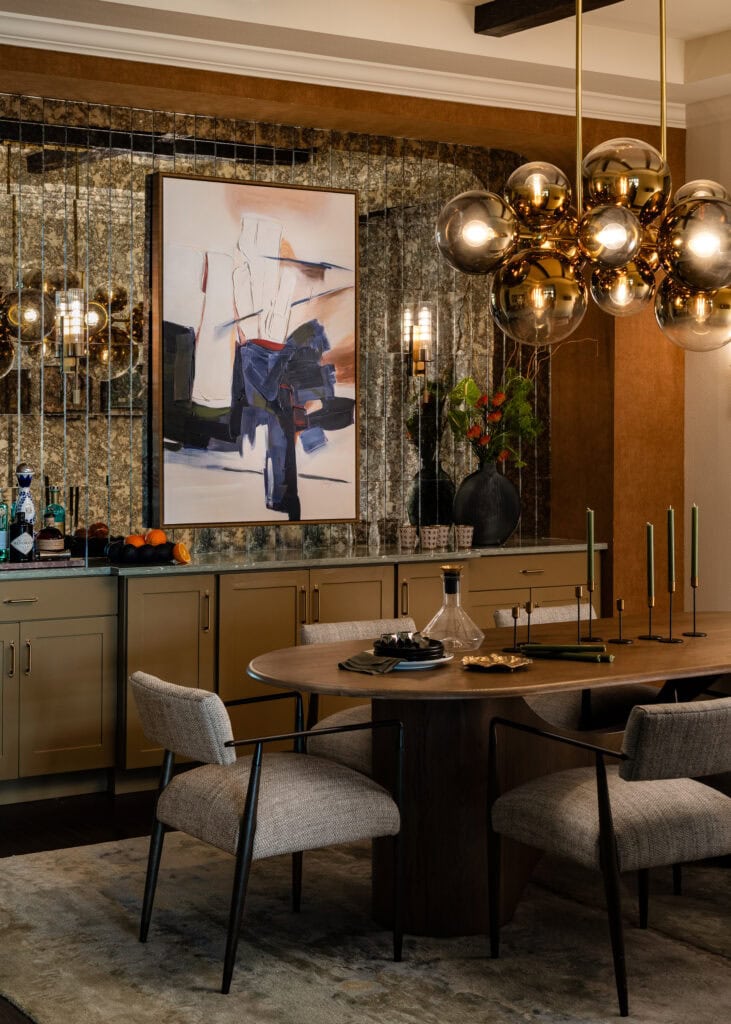 Dining area with a round wood table, bouclé chairs, a sculptural brass chandelier and an antiqued mirror wall with bold artwork, designed by HLM Designs