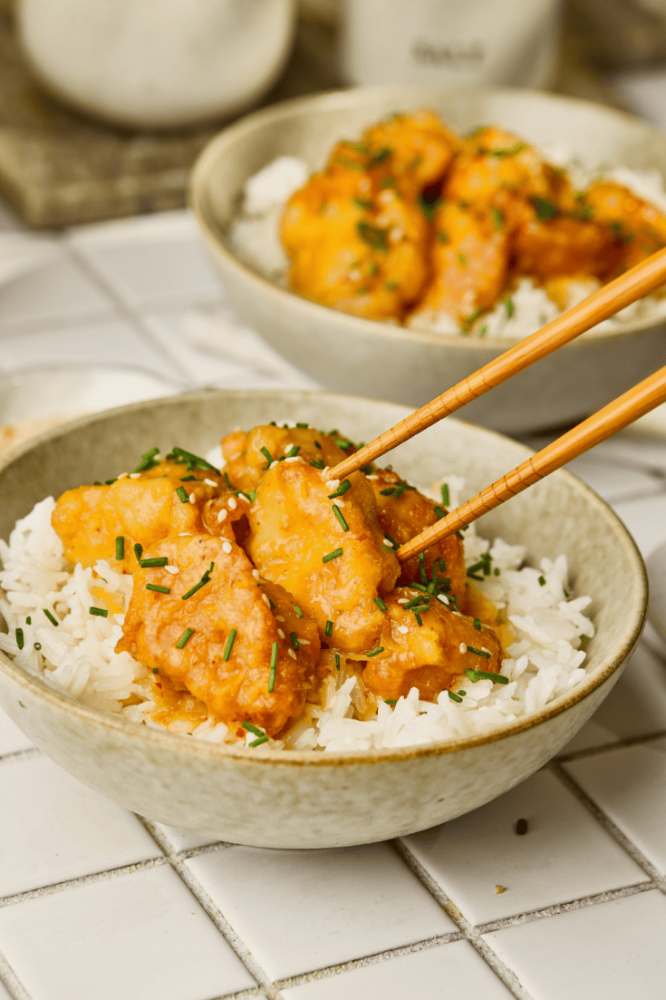 A close-up of a crispy orange chicken piece held by wooden chopsticks over a bowl of white rice.