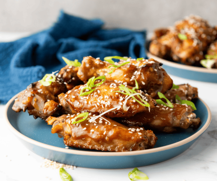A medium-angle eye-level shot of the main blue plate piled high with glazed Chinese Chicken Wings, showcasing the sesame and scallion garnish, with a second plate visible in the background against a light grey wall and blue napkin.