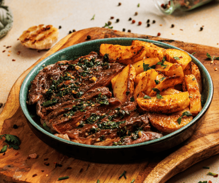 Sliced steak and garlic fries in a bowl on a rustic wood background with roasted garlic and peppercorns.