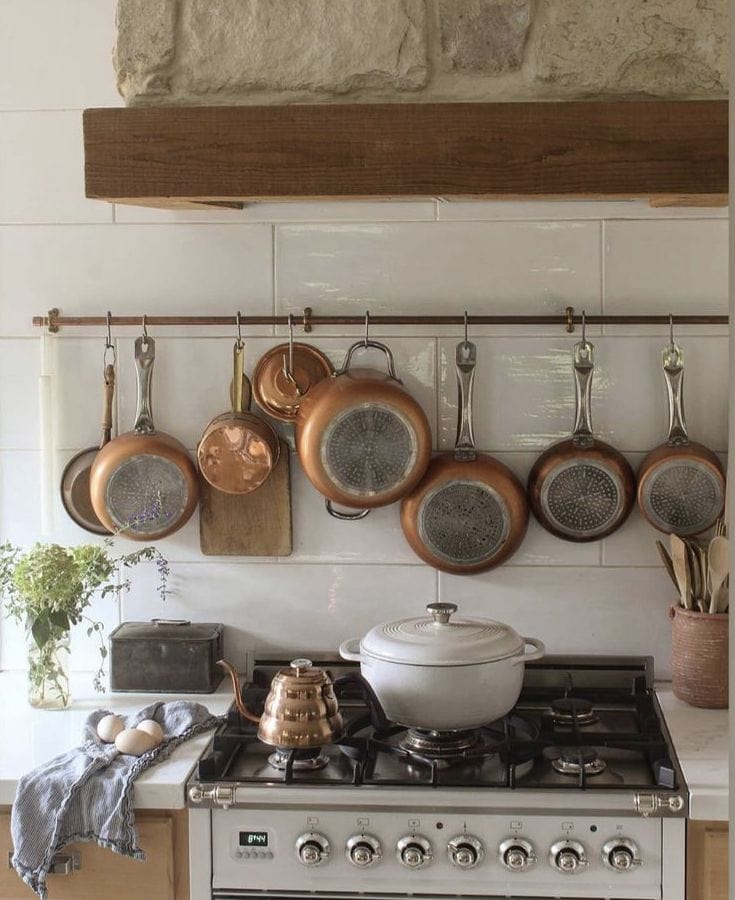 Warm kitchen with wooden cabinets, white stove, and marble counters styled with copper pots, a gold tea kettle, and a white cooking pot — cozy top of stove decor ideas from By Paige Nicole.