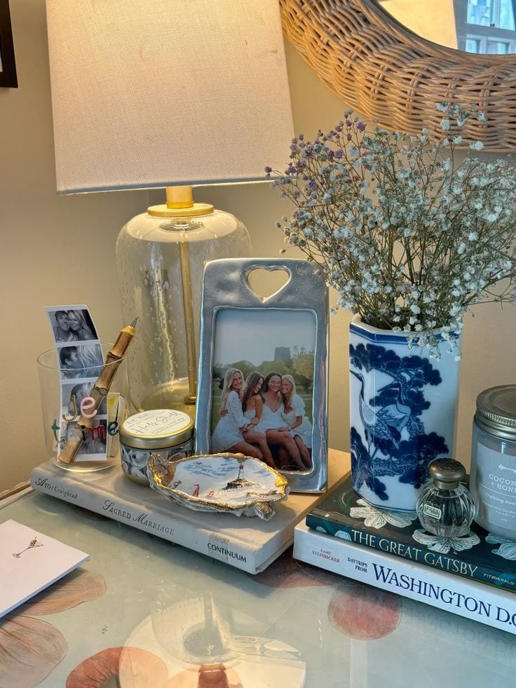 Nightstand styled with a stacked book set, lamp, jewelry dish, and framed photo of friends at the beach — relaxed blue and white coastal bedside table decor ideas from By Paige Nicole.