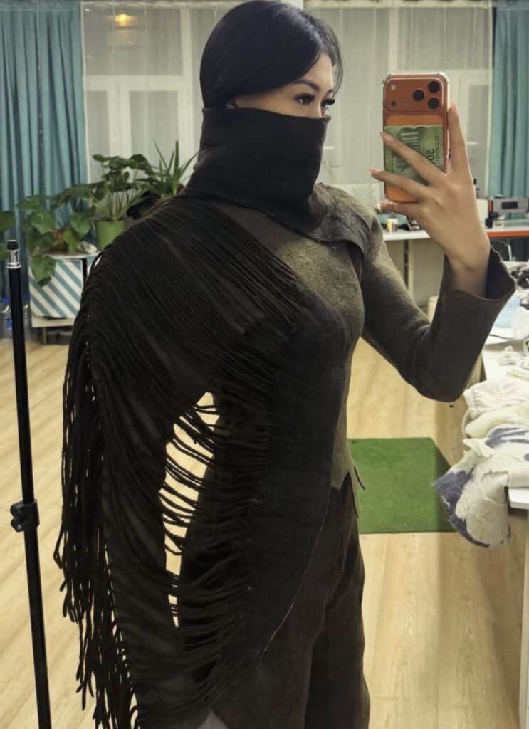 A close-up of a model wearing a dark, felted wool top by Meerim Naizabekova, featuring long, organic fringe and a built-in face covering as part of the "Balance" collection.