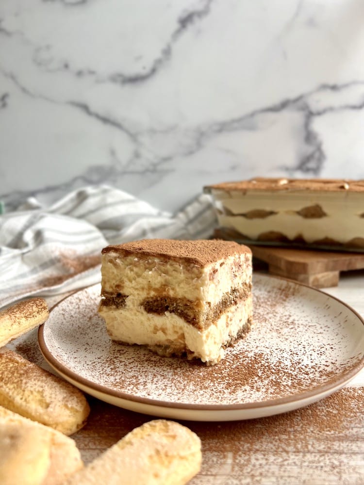Guinness and Irish Cream Tiramisu 1 A slice of Guinness and Irish Cream Tiramisu on a plate dusted with cocoa powder with a pan of tiramisu in the background