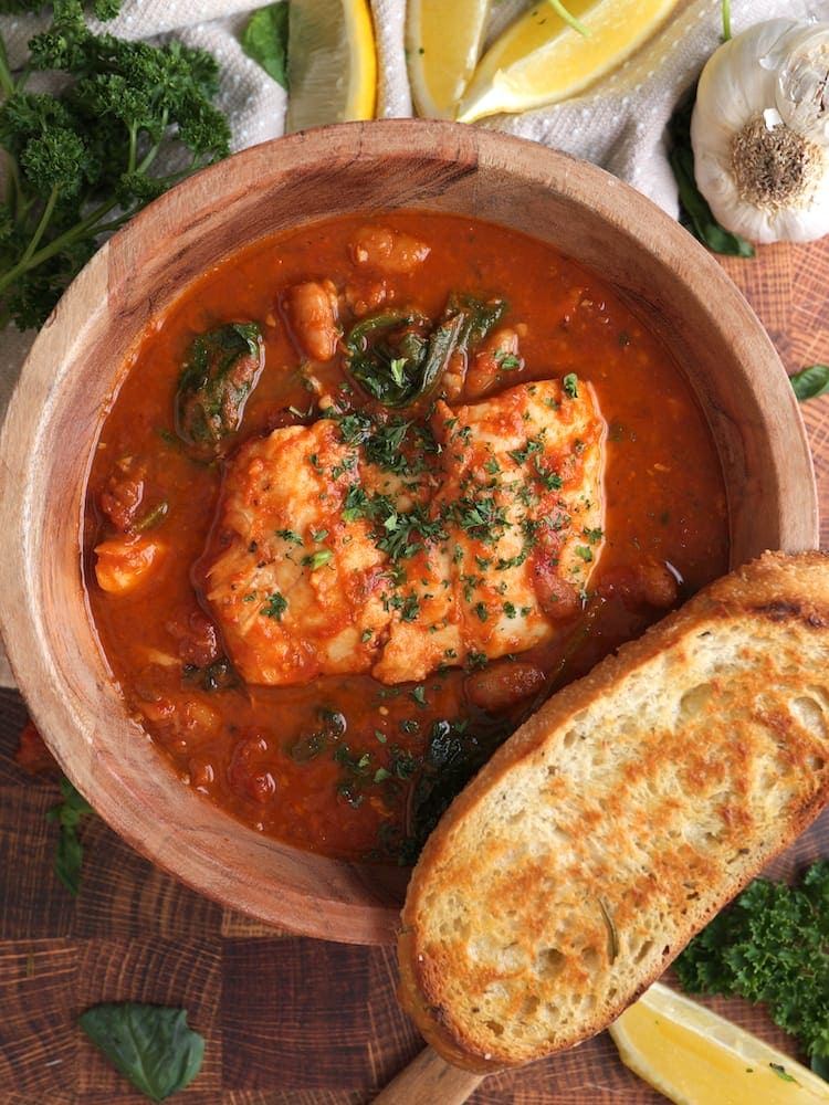 Tuscan Fish Stew 1 Tuscan Fish Stew in a wooden bowl with a slice of toasted bread on the rim of the bowl and the bowl surrounded by lemon wedges, fresh parsley, and a head of garlic