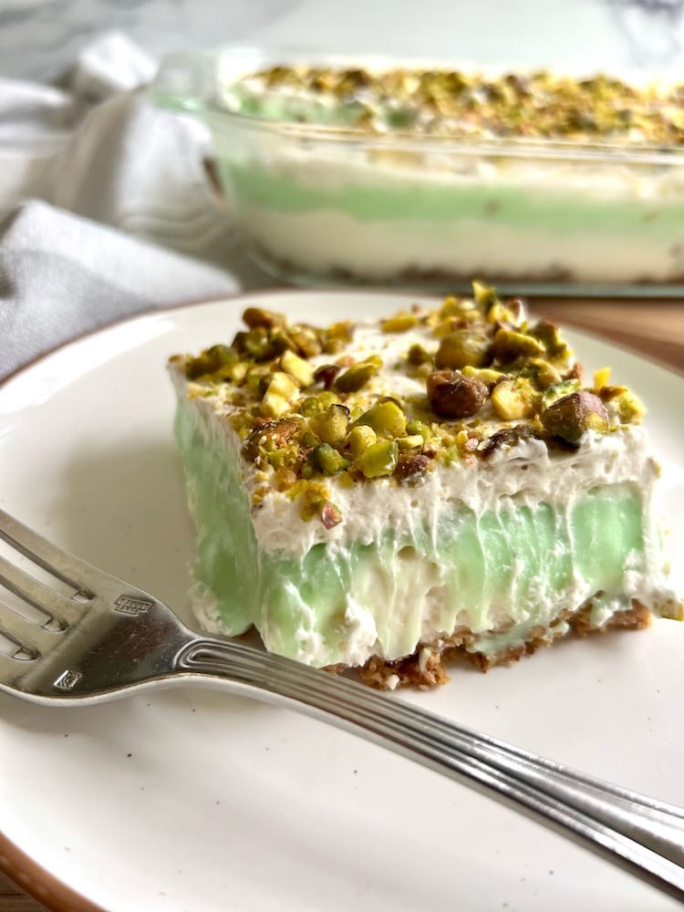 Grammie's Layered PIstachio Dessert 1 A slice of Grammie's Layered Pistachio Dessert on a plate with fork, and a pan of Grammie's Layered Pistachio Dessert in the background.
