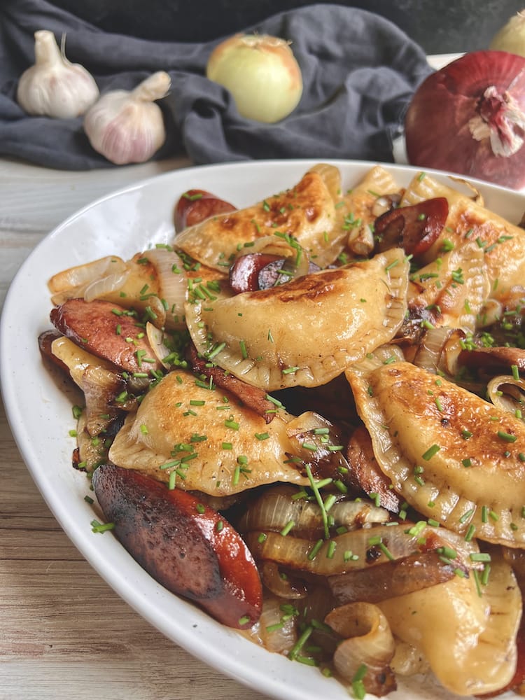 Griddled Pierogi & Kielbasa 1 WEB Griddled Pierogi and Kielbasa in a large white serving dish with fresh garlic and onion in the background