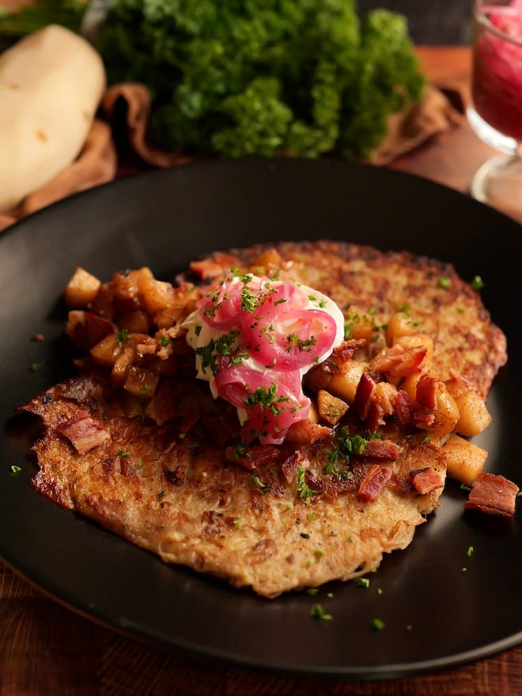 Sweet & Salty Latkes - 4 WEB Sweet and Salty Latkes topped with pickled red onion, caramelized apples, bacon, and sour cream on a black plate