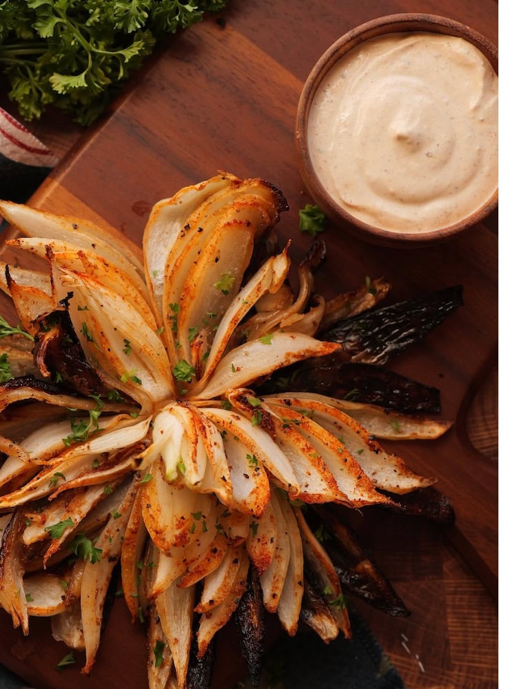 Grilled Blooming Onion 2 WEB A Grilled Blooming Onion on a cutting board with a ramekin of dipping sauce next to it