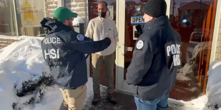 Two officers in HSI jackets speak with a man at a store entrance on a snowy sidewalk, near a glass door with an 'OPEN' sign visible.