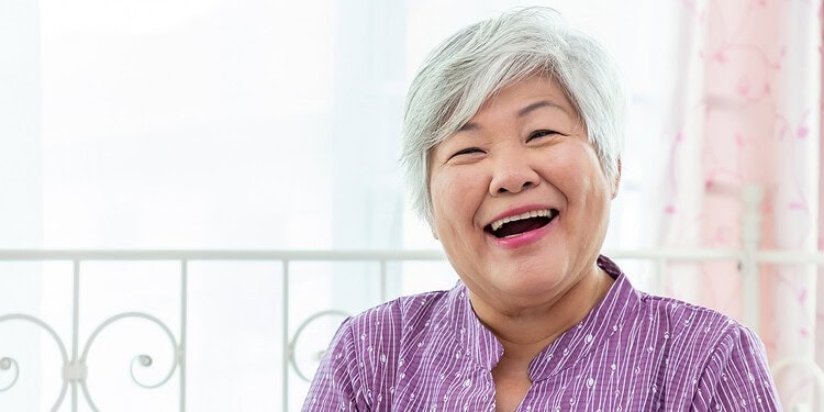 Joyful elderly woman with gray hair laughing, bright background, expressing happiness and positive emotions in a home setting.
