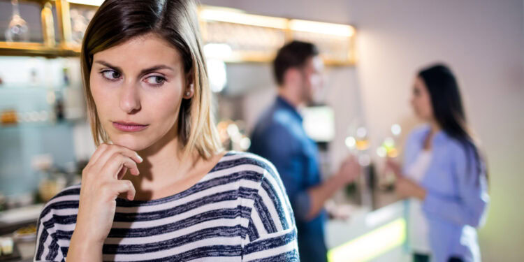 Reflecting woman with pensive expression in a social setting, two people talking in the background.