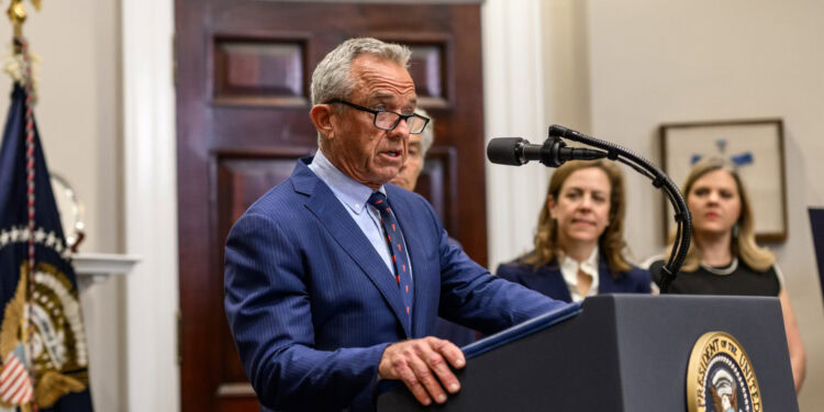 Elderly man in a blue suit speaking at a podium with a microphone during a formal event or press conference.