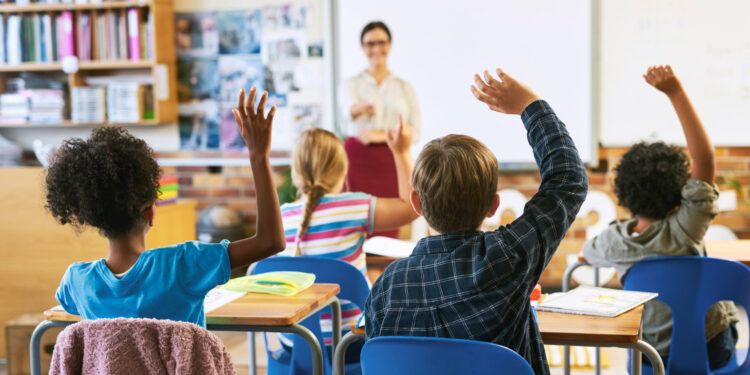 Children in classroom raising hands to answer teacher's question.