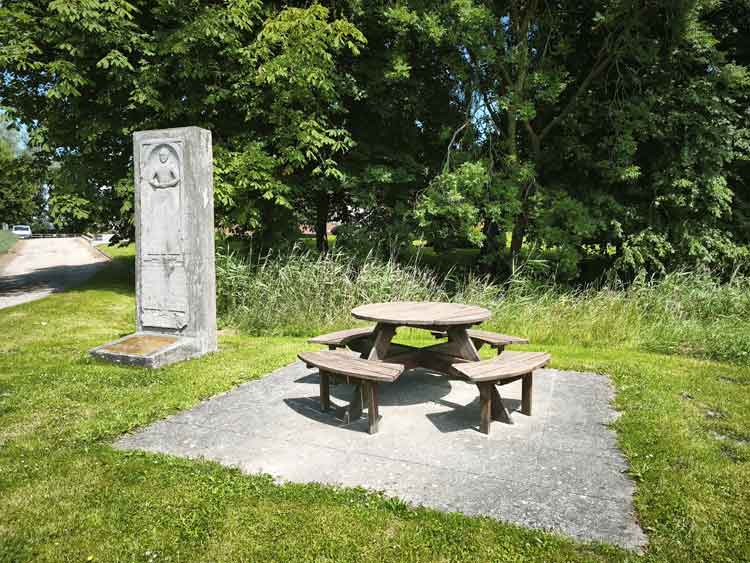 Auf einer Wiese steht ein Gedenkstein. Eine Sitzgruppe aus Holz daneben lädt zur Pause ein. Es Bäume stehen dahinter.