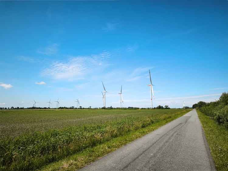 Eine gerade lange Straße, ein Blick auf Felder und einen Windradpark.