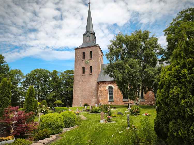 Ein Blick auf die Kirche in Oldenswort. Die Kirche ist ein typisch friesischer Backsteinbau. Der Friedhof ist sehr gepflegt und dicht bepflanzt. 