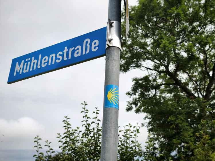 Ein Straßenschild mit einem Aufkleber der Jakobsmuschel, dem Wegweiser der Pilgernden.