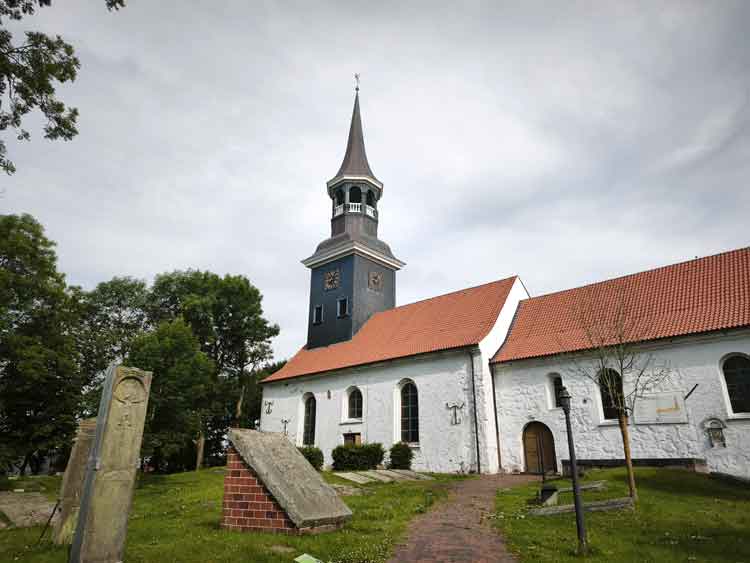 Ein Blick auf die Lundener Kirche. Sie ist weiß mit rotem Dach und hat einen grauen Kirchturm. Davor befinden sich die Steine des Geschlechterfriedhofs.