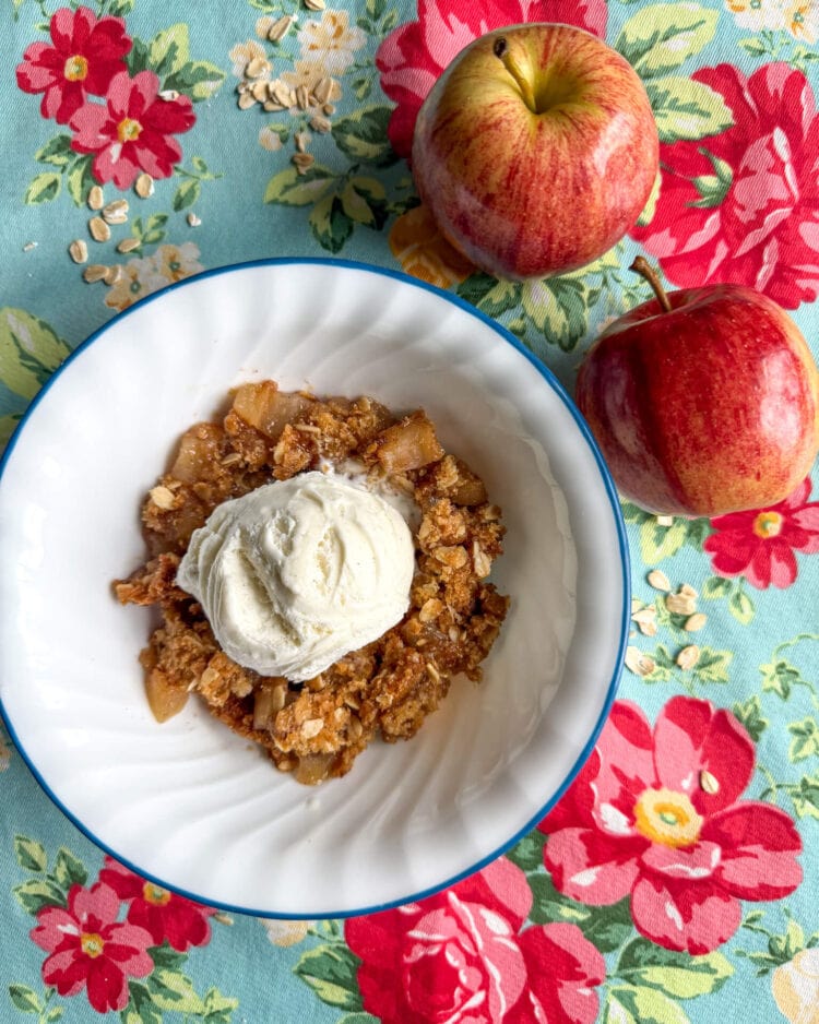 Bowl of fresh milled flour apple crisp on a bright colored kitchen placemat with two apples on the side.